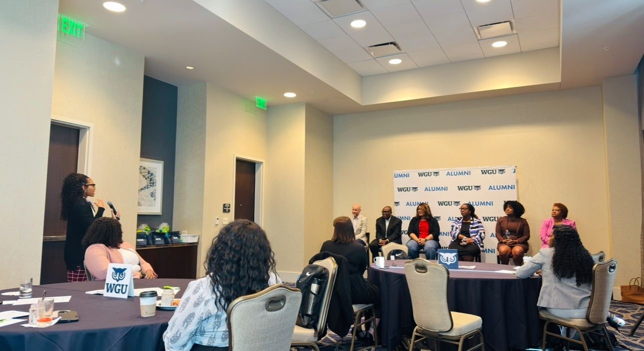 A woman speaks into a microphone at a WGU Alumni event, with a panel of five people seated on a stage and an audience at round tables.